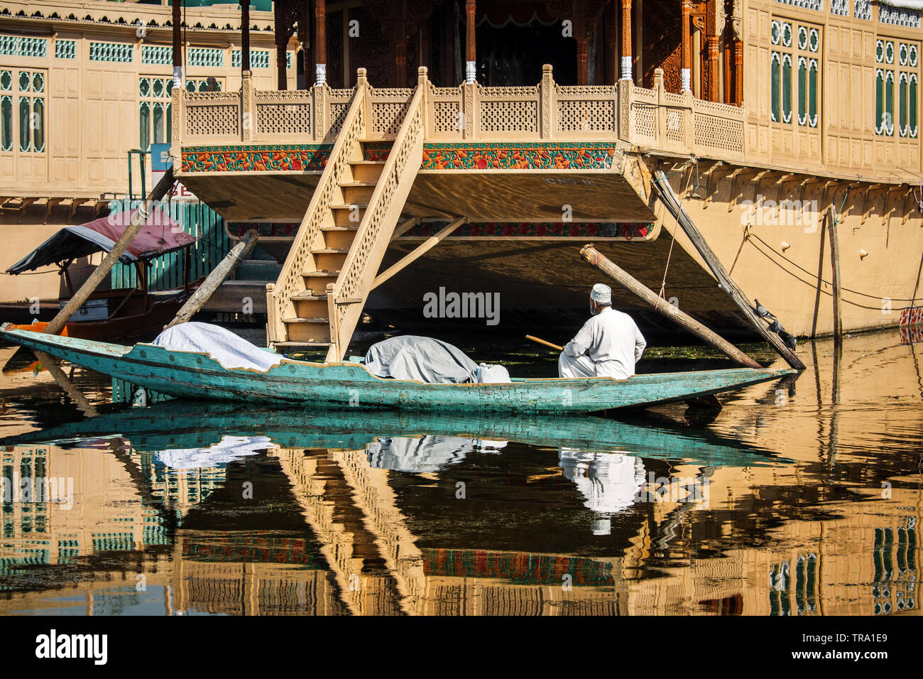 Muslimischen Mann mit an der Rückseite der Kamera in einem langen Boot Rudern neben wunderschön verzierten Hausboote auf dem berühmten Dal Lake von Srinagar in Kaschmir Valley Stockfoto