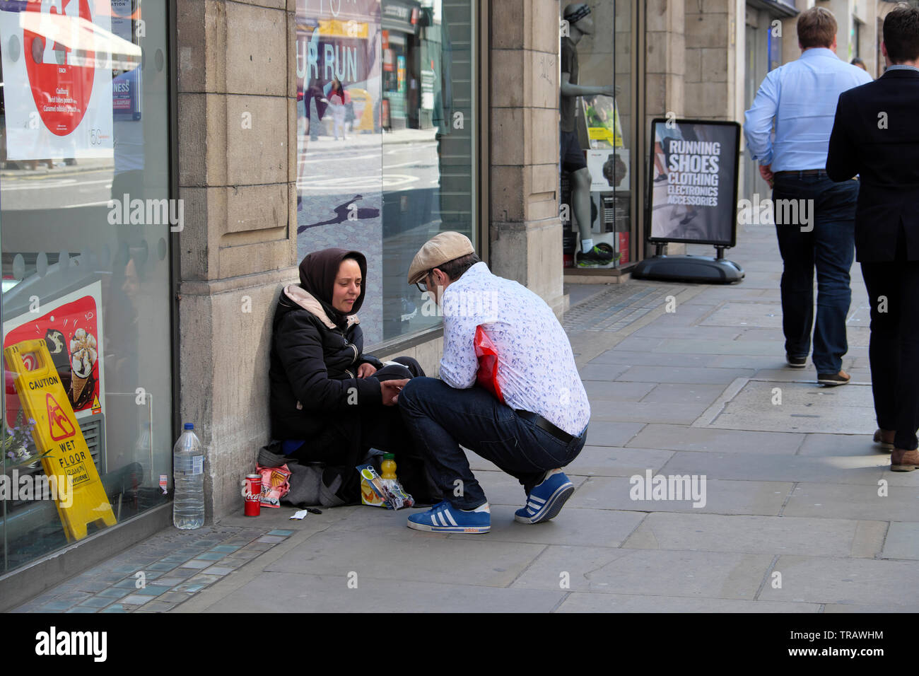 Eine Art Mann bietet Speisen und Getränke für eine obdachlose Frau betteln auf der Straße vor einem Supermarkt mit Menschen zu Fuß durch London England UK KATHY DEWITT Stockfoto