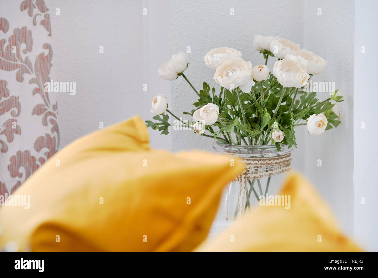 Gelbe Kissen auf der Couch im Wohnzimmer, am wunderschönen Blumen auf einem dünnen Stamm in Glasvase weißen Ranunkeln in der Nähe der Wall niedlich Dekoration Zuhause Stockfoto