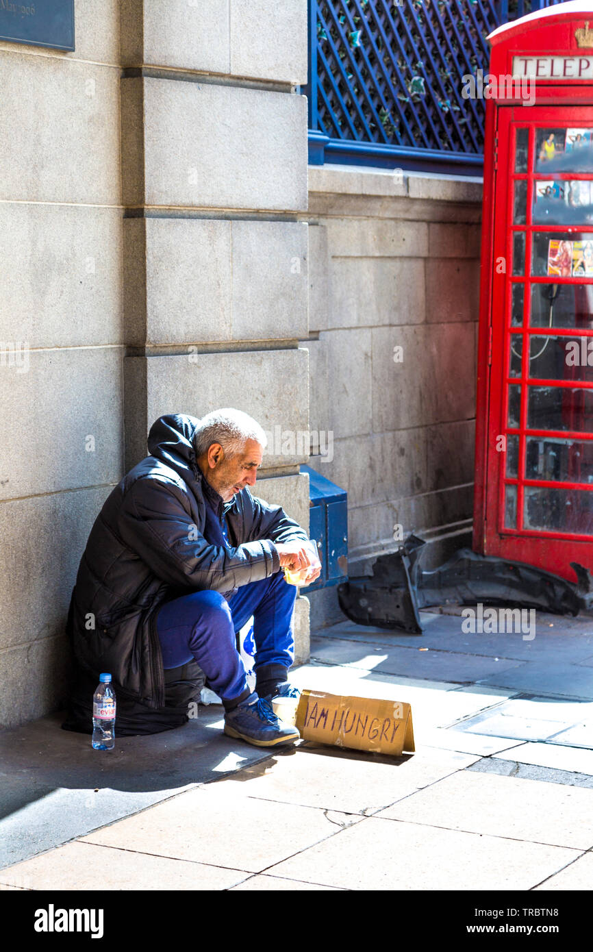 Hungriger Obdachloser mit 'Ich habe Hunger'-Schild auf dem Bürgersteig sitzend und essend vor dem Ritz, Green Park, London, UK Stockfoto