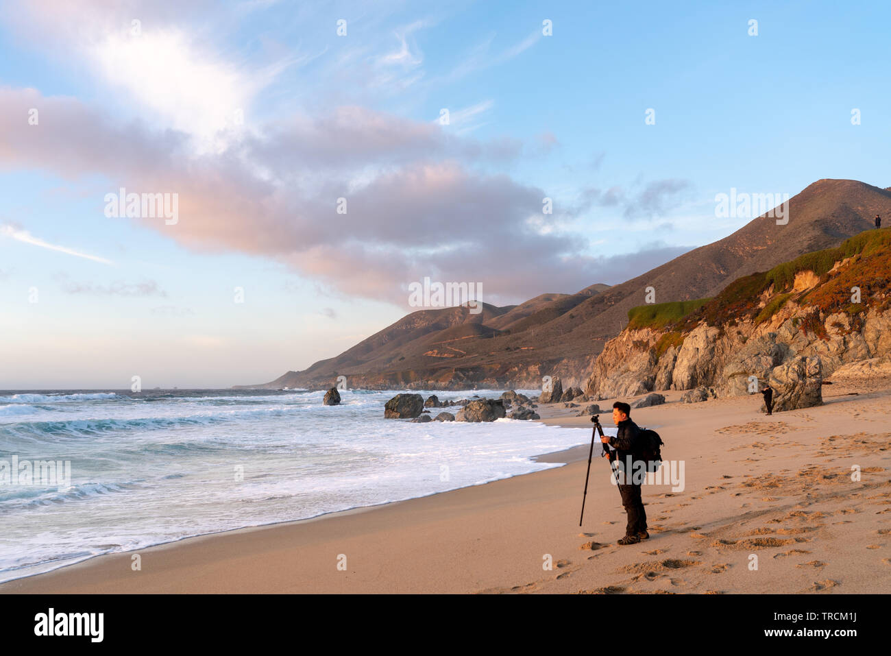 Asiatischer Mann setzt Stativ auf einem Sandstrand an der Küste von Big Sur, Kalifornien während des Sonnenuntergangs. Stockfoto