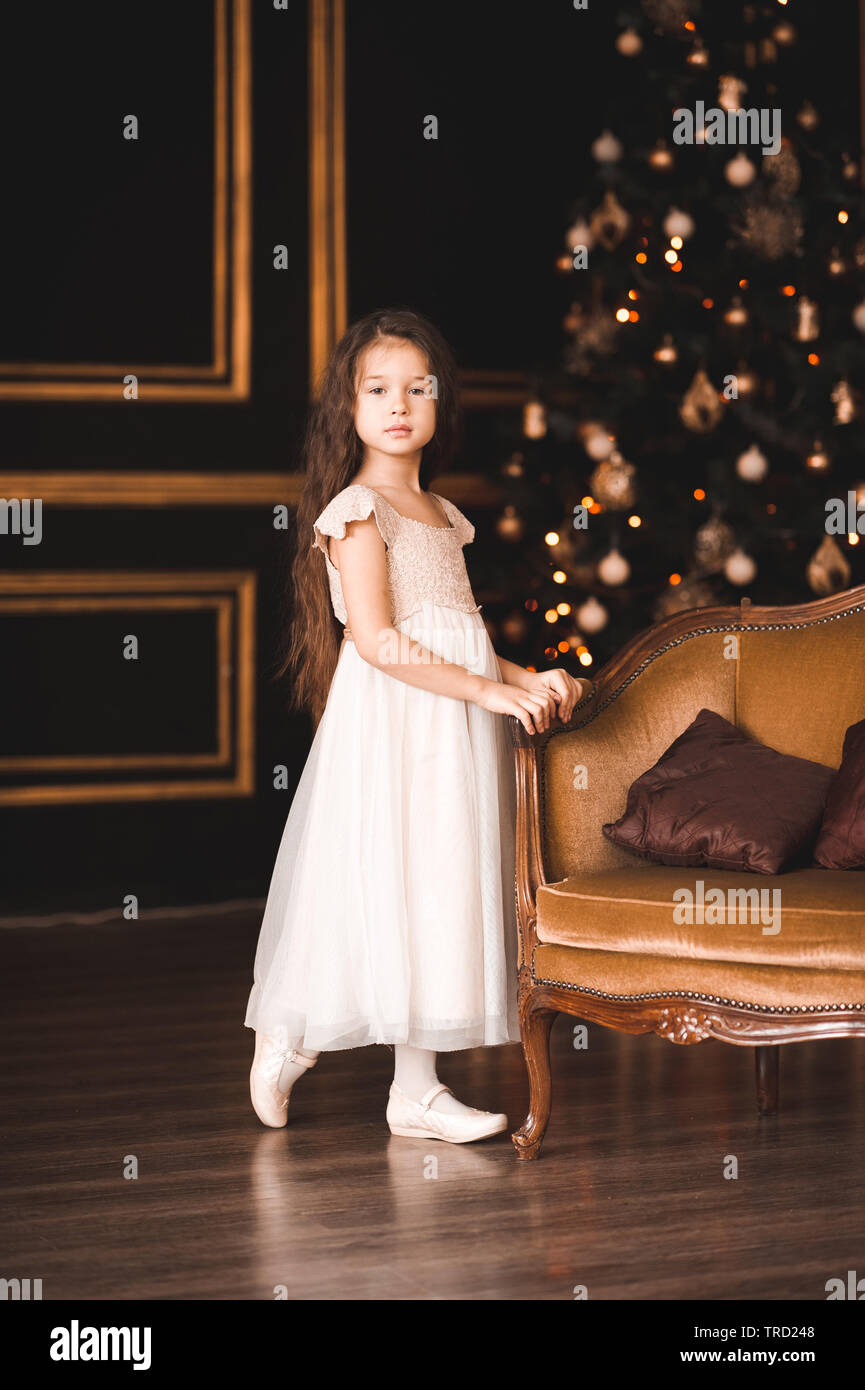 Cute Baby Mädchen 4-5 Jahre alten tragen elegantes weißes Kleid über Weihnachten Lichter im Raum darstellen. Mit Blick auf die Kamera. Urlaubszeit. Stockfoto