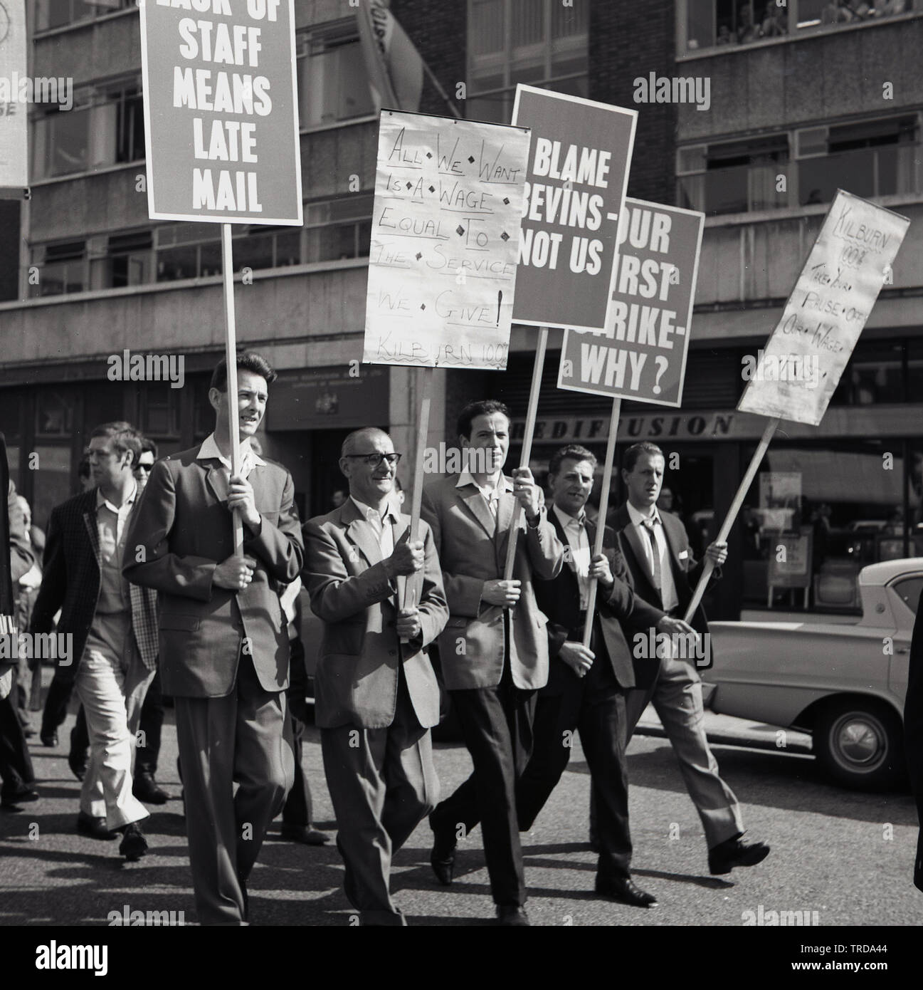 1960s, historische, streikende Royal Mail Postarbeiter, die mit Plakaten durch das Zentrum Londons laufen, eine davon..."Unser erster Streik Warum? Die Postboten der Zweigstelle von Kilburn protestierten gegen ihre niedrigen Löhne und forderten einen gescheiterten Lohnvertrag, mit besonderer Angst vor Ernest Bevin, dem Mitbegründer des Unternehmens, Im Januar 1922 - und ehemaliger Generalsekretär - der Transport and General Workers Union (TGWU), die zu Großbritanniens größter Gewerkschaft geworden war. Stockfoto