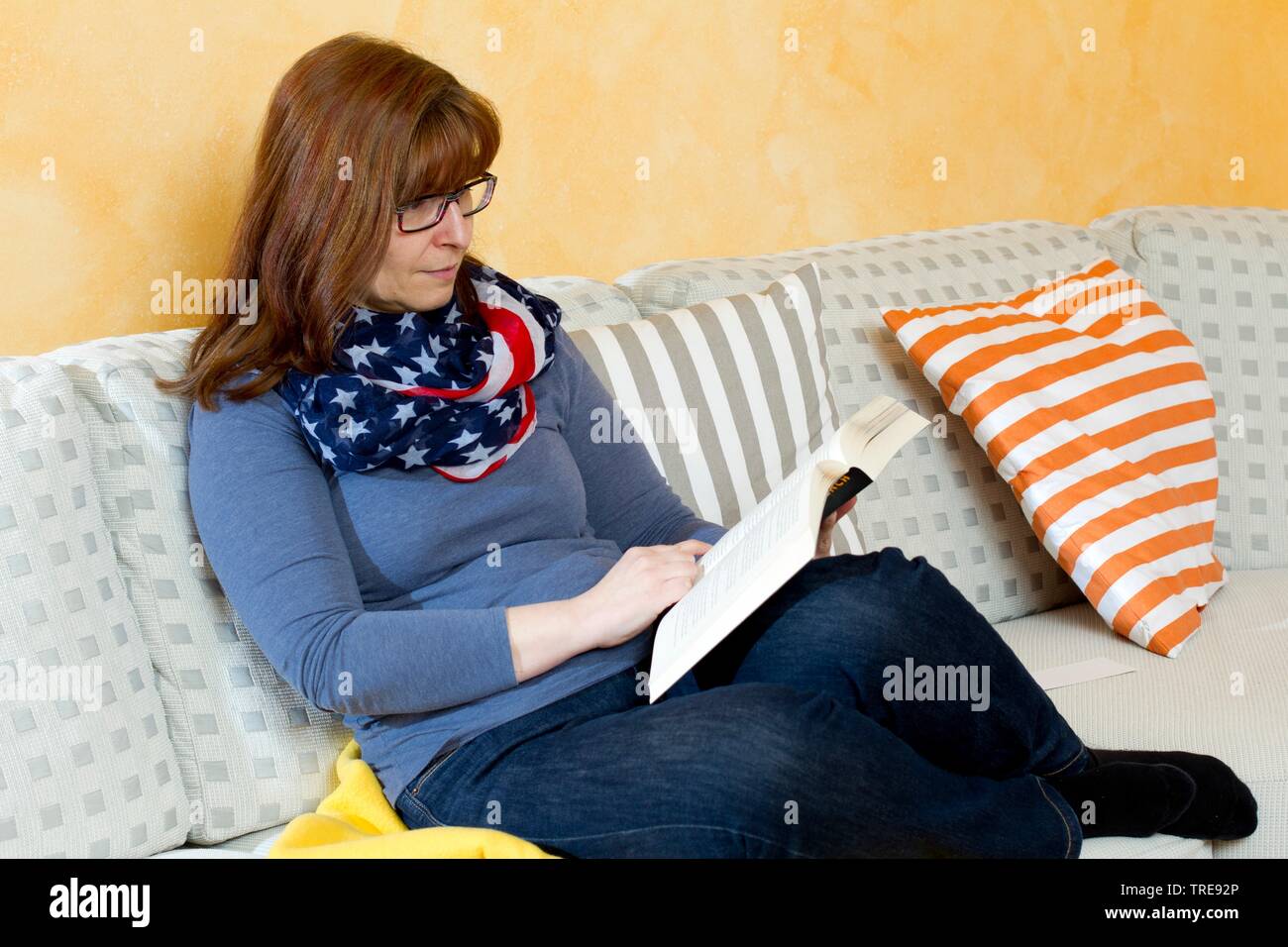 Frau mittleren Alters mit roten Haaren Brille sitzt auf einem Sofa in einem Buch lesend Stockfoto