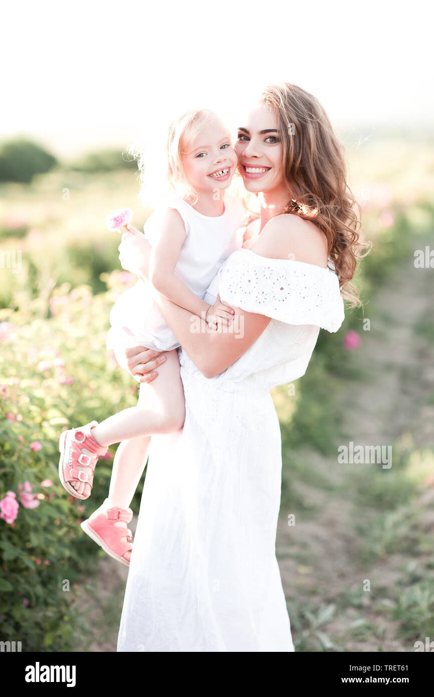 Lächelnde Mutter Holding kleinen Mädchen 4-5 Jahre alten tragen ähnliche weiße Kleider stehen in der Wiese. Mit Blick auf die Kamera. Die Mutterschaft. Mutterschaft. Familie Stockfoto