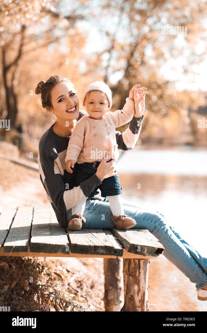 Lächelnde Mutter Holding baby girl 1 Jahr alt tragen aus Gewirken legere Kleidung im Herbst Park sitzen. Mit Blick auf die Kamera. Die Mutterschaft. Stockfoto