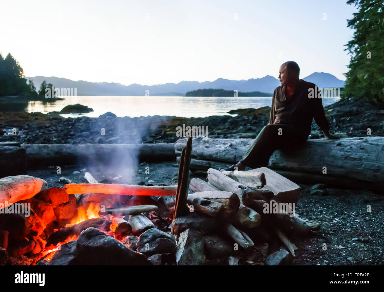 Mann sitzt auf Strand am Lagerfeuer in der Wildnis, Broken Group Islands, Barkley Sound, Pacific Rim National Park, Kanada. Selektiver Fokus auf Feuer. Stockfoto