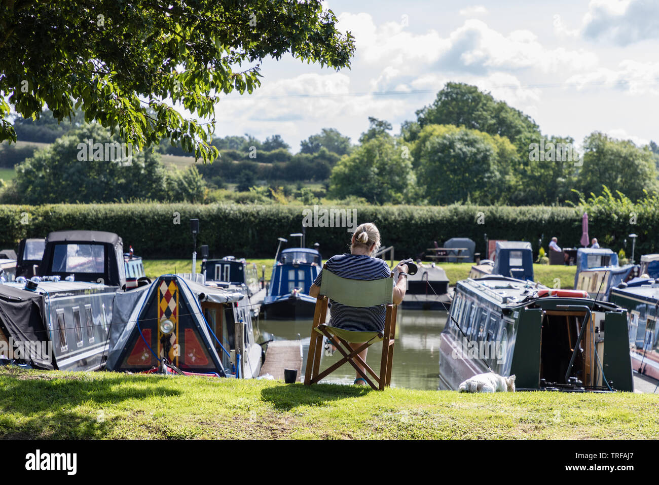 Crick Marina, Northamptonshire, Großbritannien: Eine Frau sitzt in einem klappbaren Stuhl, während sie einen Hund an der Leine hält. Vor ihr liegen Kanalengboote. Stockfoto