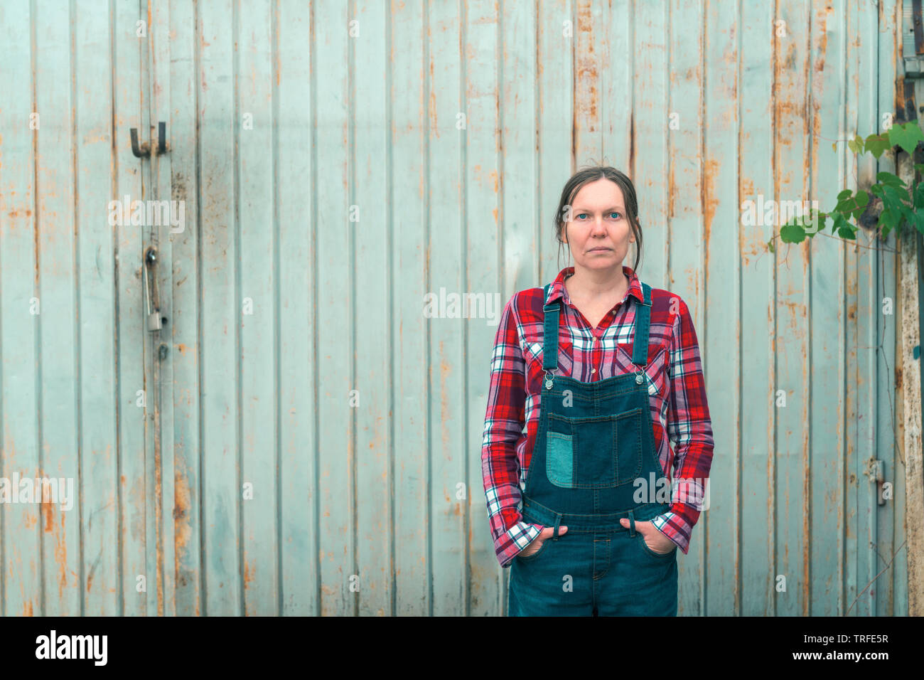 Porträt der schönen Frau Bauer vor Bauernhof Schuppen. Zuversichtlich landwirtschaftlichen Arbeitnehmers tragen Plaid Shirt und Jeans mit Hosenträgern sucht ein Stockfoto