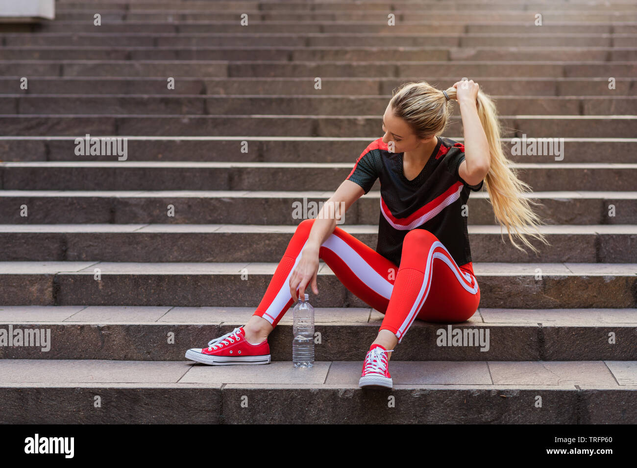 Junge Frau in Active Wear sitzen auf Treppen mit Wasserflasche. sie trägt rote Übung Hosen und rote Schuhe. Stockfoto