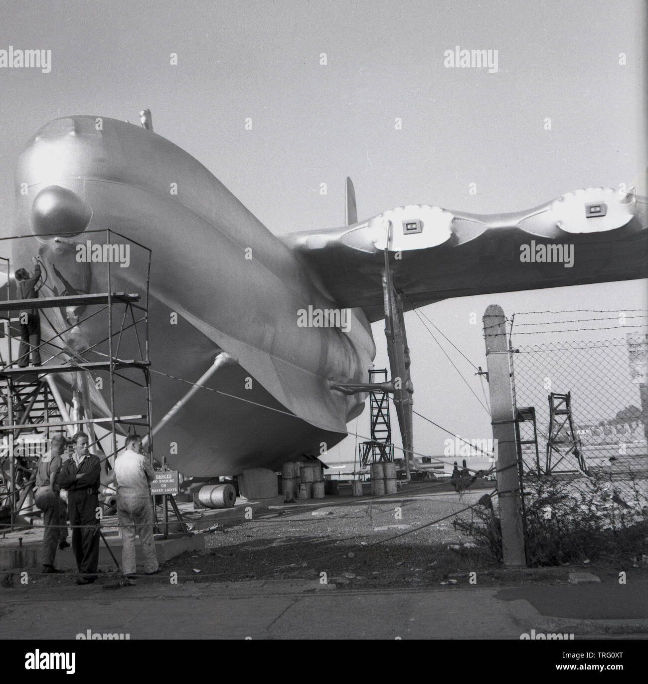 1950, historische, Arbeitnehmer in Overalls außerhalb im industriellen Bootswerft arbeiten auf eine volle Größe 'Modell' Wasserflugzeug, eventuell für die Verwendung als stgae prop in einem Spielfilm. oder den Film. Ein Arbeiter auf Gerüst mit einem lackspray auf der Vorderseite des Flugzeugs. Stockfoto