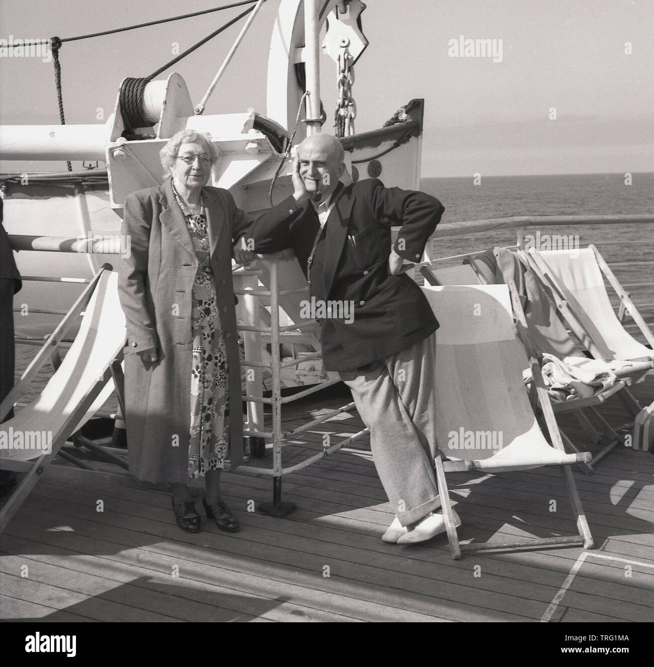 1950, historische, ein älteres Paar auf einer Kreuzfahrt stehend auf dem Deck der P&O Ocean Liner, SS Chusan. Die Frau trägt ein langes Kleid, mit ein Mantel, während der Mann mehr passend für Kreuzfahrt in einem Blazer und deck Schuhe gekleidet ist und das Rauchen einer Pfeife. Stockfoto