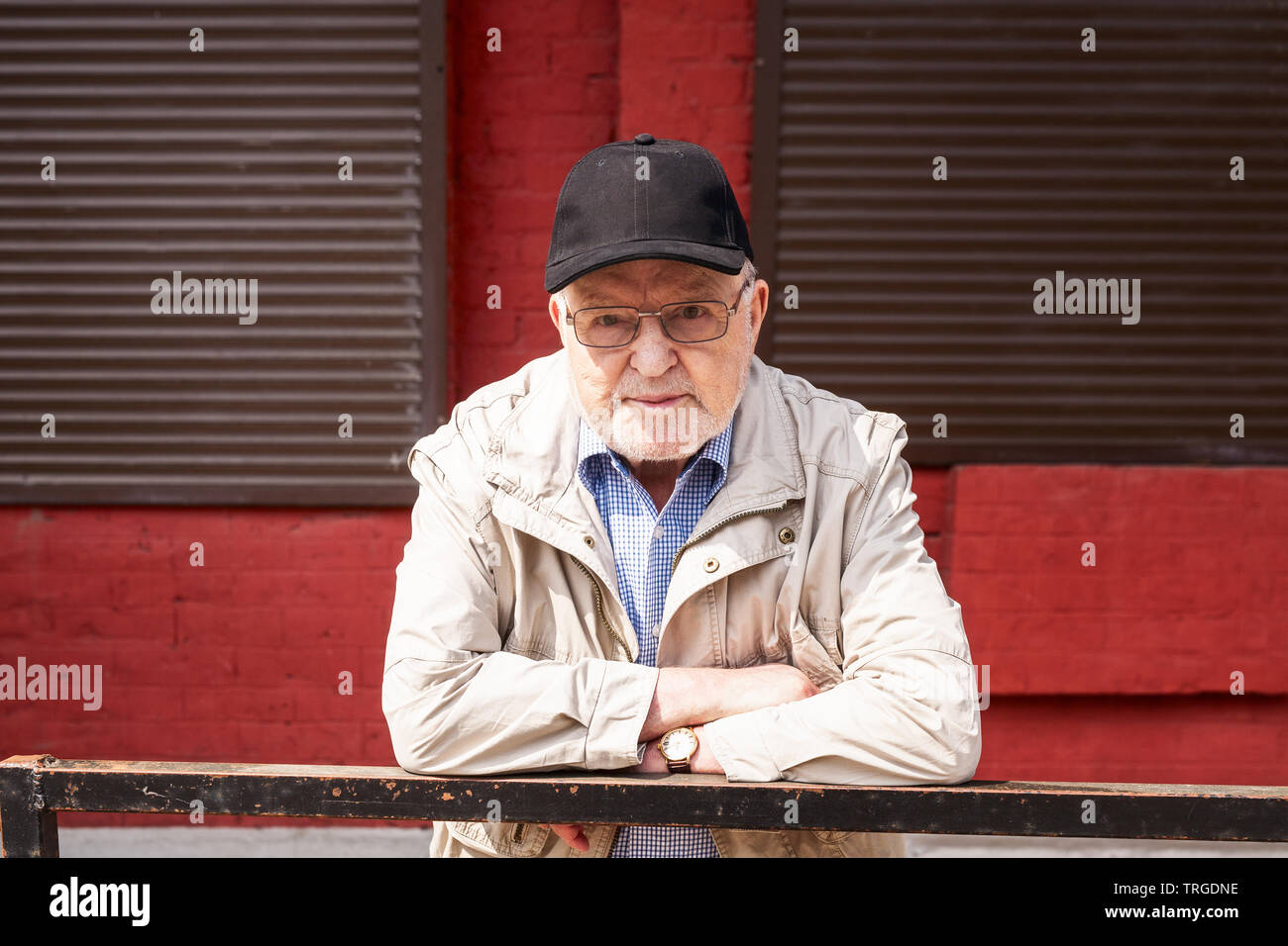 Street Portrait von alter Mann in Brillen und Kappe im Freien. Senior Posieren vor der Roten Wand mit Fenstern Stockfoto