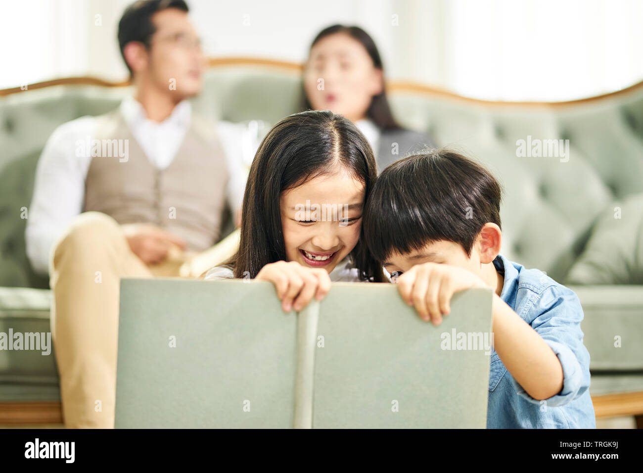 Zwei asiatische Kinder Bruder und Schwester auf dem Teppich lesen Buch sitzen zusammen in der Familie Wohnzimmer mit Eltern sitzen auf der Couch im Hintergrund. Stockfoto