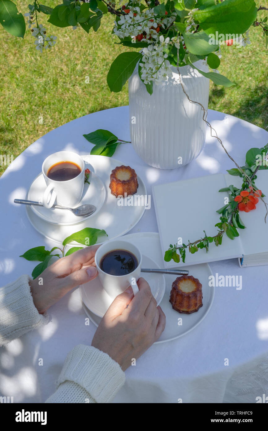 Genießen Kaffee Zeit im Garten. Serviert Tisch mit Blumen, Buch, Kaffee und Dessert im Garten im Frühling. Anordnung mit den Händen einer Frau Stockfoto