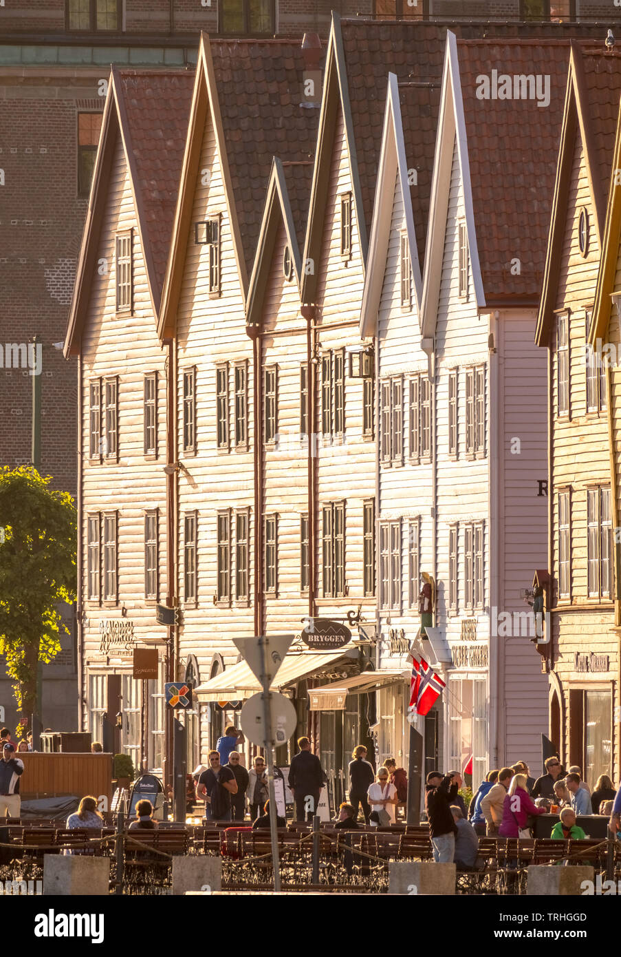 Touristen zu Fuß, andere an den Tischen auf Stühlen vor einem weißen Holzhäuser vorne mit Geschäften im Hafen von Bryggen, Bergen, Hordaland, Norwegen sitzen, Stockfoto