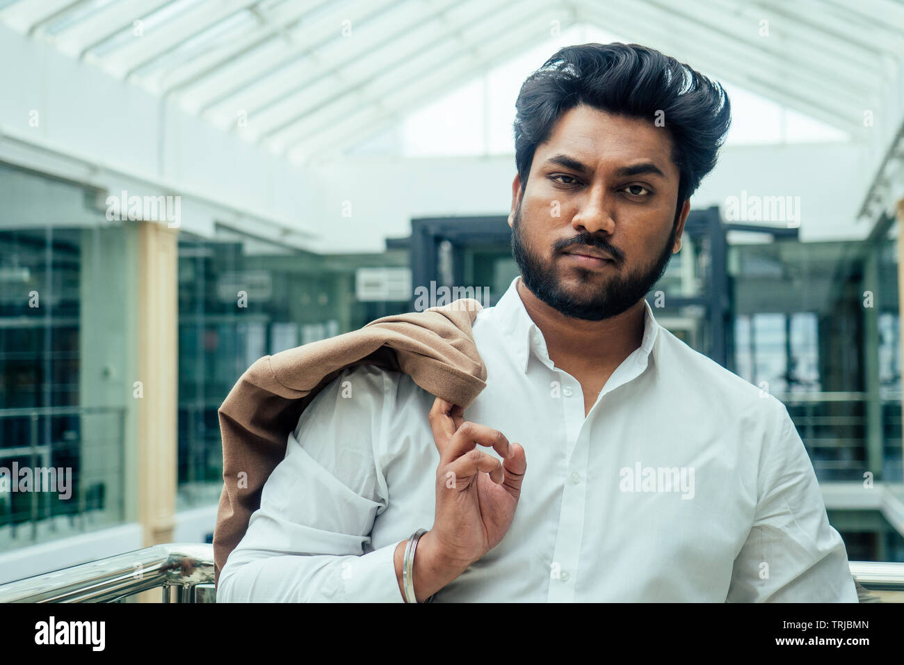 Indische Mann in einem weißen Hemd in der Hand halten, eine Braune Jacke in einem modernen Büro mit Glasfenstern Shopping Mall posing Stockfoto