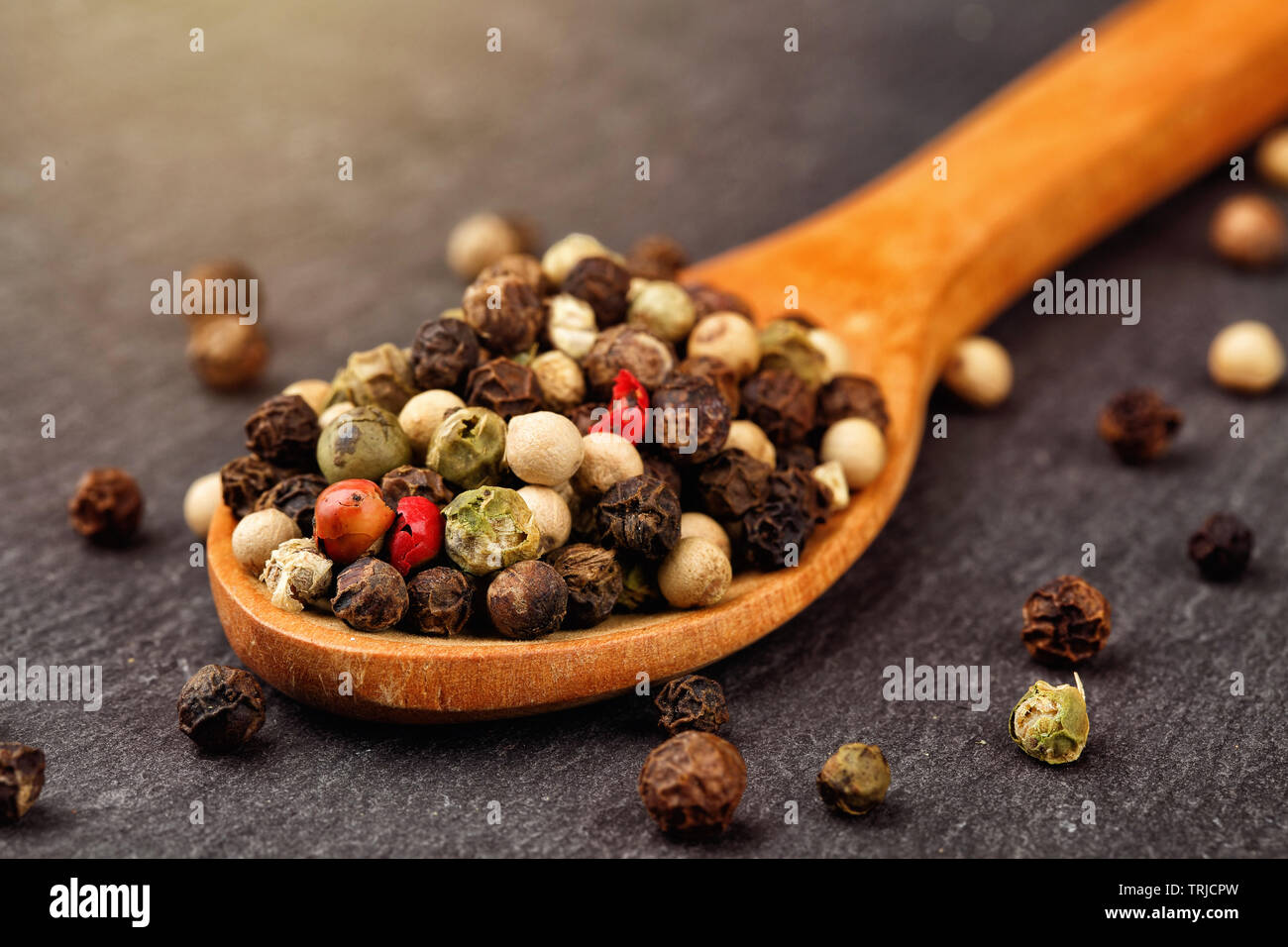 Makroaufnahme von Schwarz, Grün, Weiß und Rot Pfeffer Mix in einem Löffel aus Holz auf einem dunklen Stein Küche mit Sun haze Licht Stockfoto