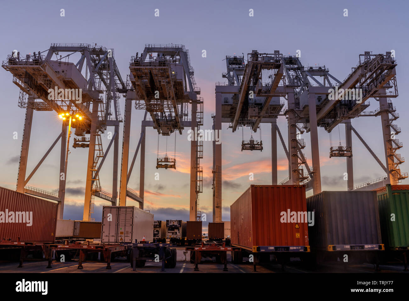 Shipping Container Krane und Lkws mit Sonnenuntergang Himmel im Hafen von Oakland. Stockfoto