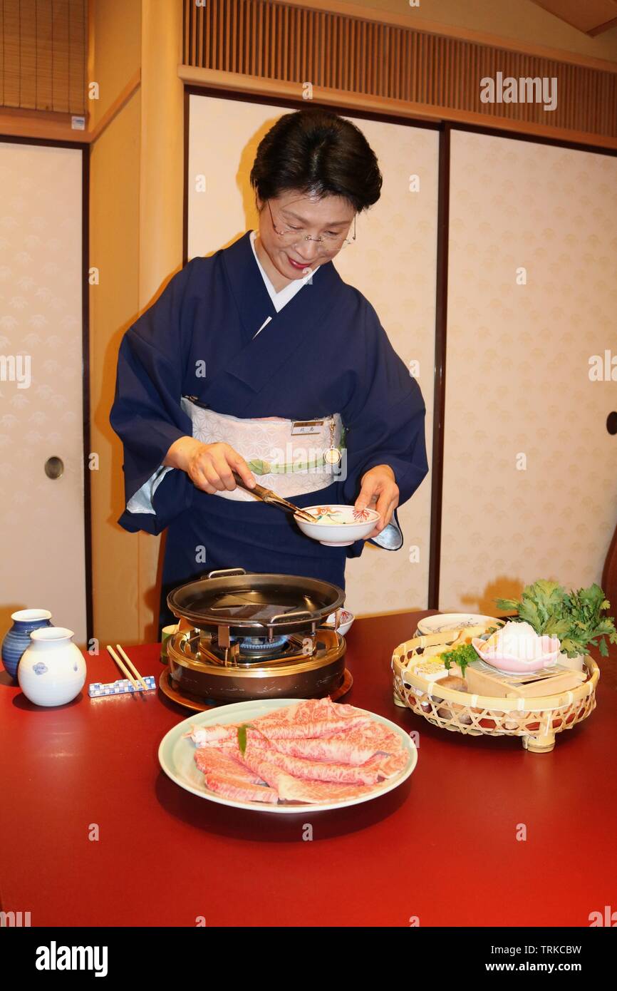 Japanische Dame in Marine kimono rühren Eier über einem kleinen Gasbrenner mit Platte von Kobe Rindfleisch vor Stockfoto