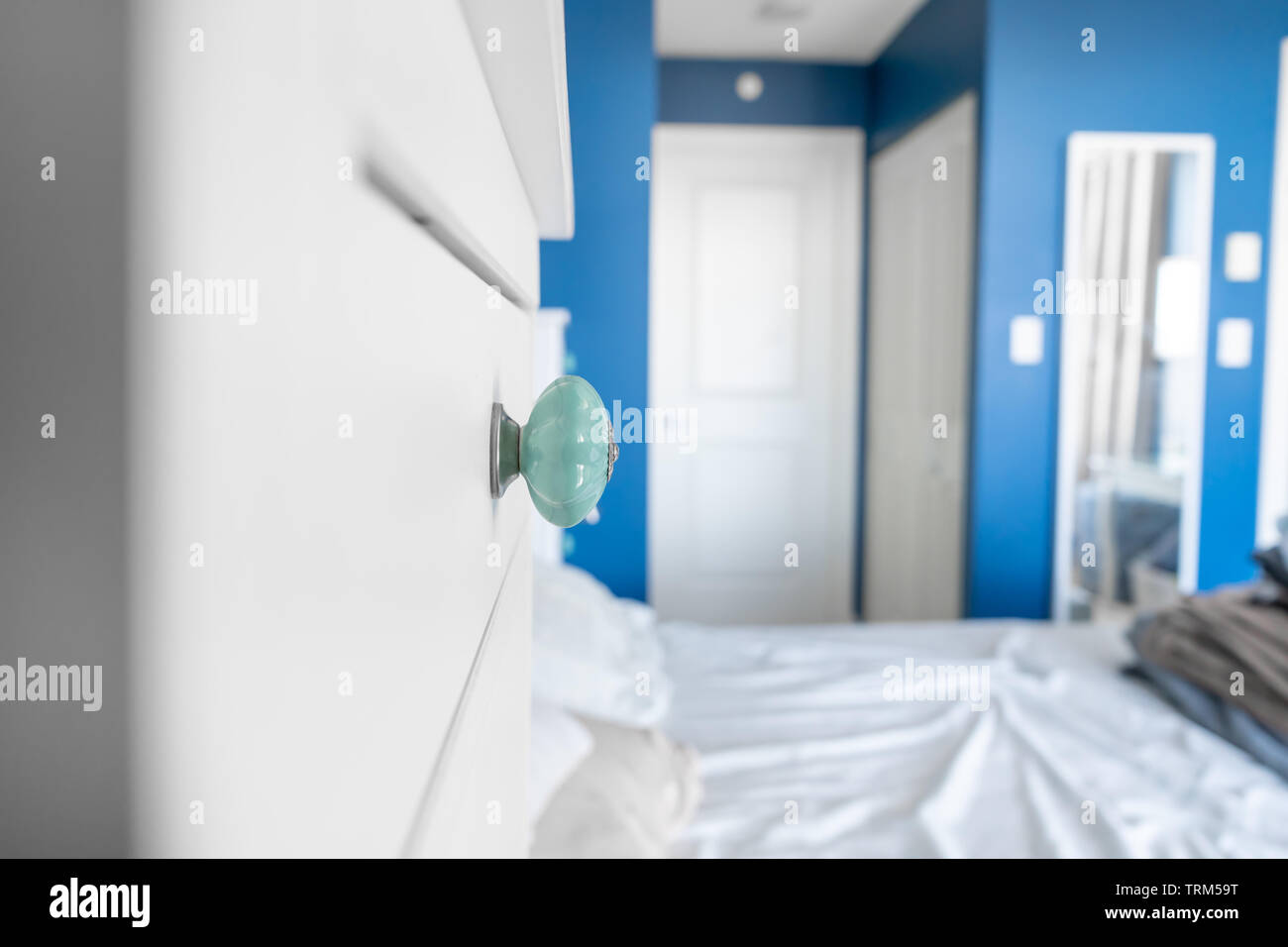Blick in ein Schlafzimmer, einen begehbaren Kleiderschrank, eine Kommode Knopf, blau gestrichenen Wänden, weißen Akzenten und ein Bett ohne Decken, mit unordentlichen, zerknitterten Bettlaken. Stockfoto