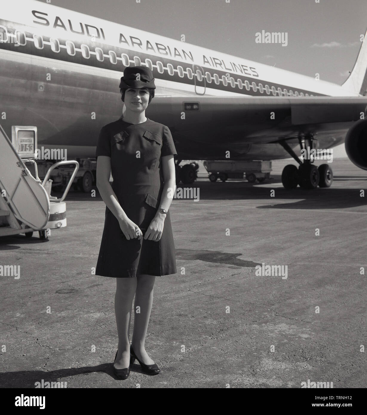 1960, historische, Uniformierte weibliche Air Hostess außerhalb einer Saudi Arabian Airlines Jet stehend, Flughafen Heathrow, London, England, UK. Stockfoto