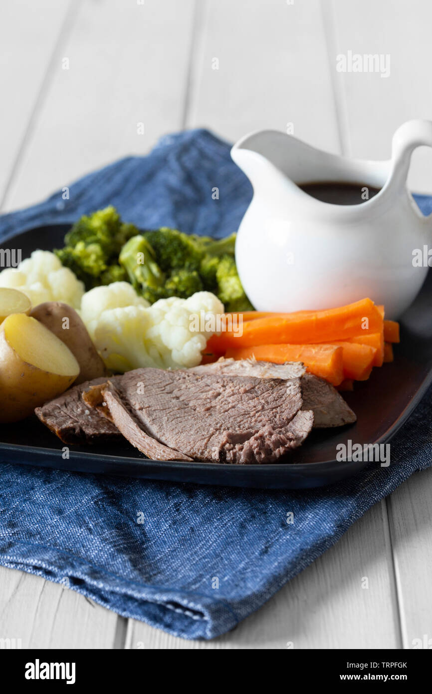 Lammbraten Sonntag Mittagessen Abendessen mit Gemüse auf einem tjeukemeer schwarze Platte, mit Soße Krug, auf einem blauen Küchenhandtuch. Weiß Holz Hintergrund Stockfoto