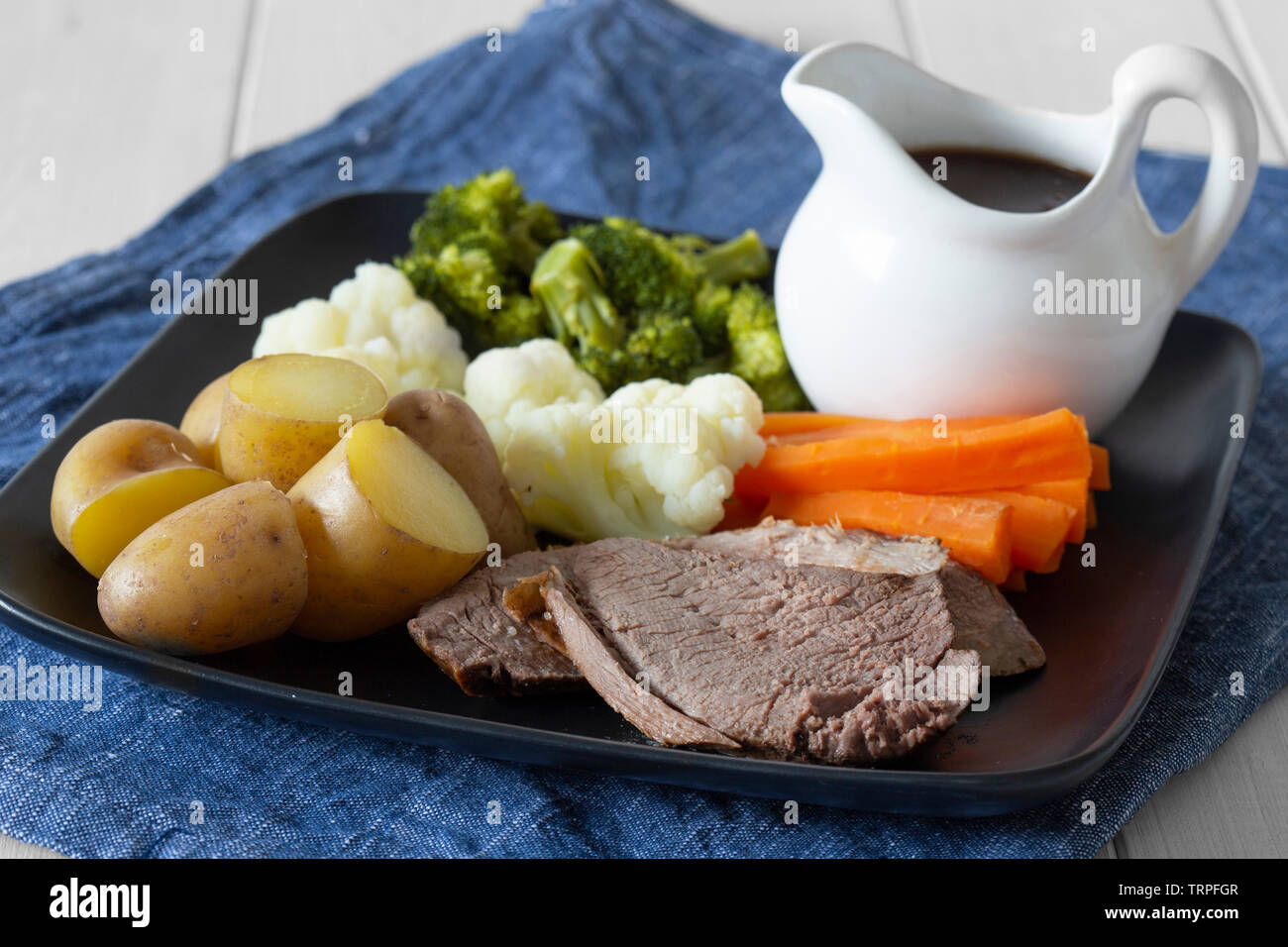 Lammbraten Sonntag Mittagessen Abendessen mit Gemüse auf einem tjeukemeer schwarze Platte, mit Soße Krug, auf einem blauen Küchenhandtuch. Weiß Holz Hintergrund Stockfoto