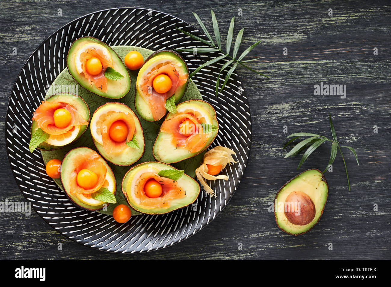 Avocado Boote mit geräuchertem Lachs, Zitrone, Physalis und Minzeblatt, Ansicht von oben auf dunkel strukturierten Hintergrund Stockfoto