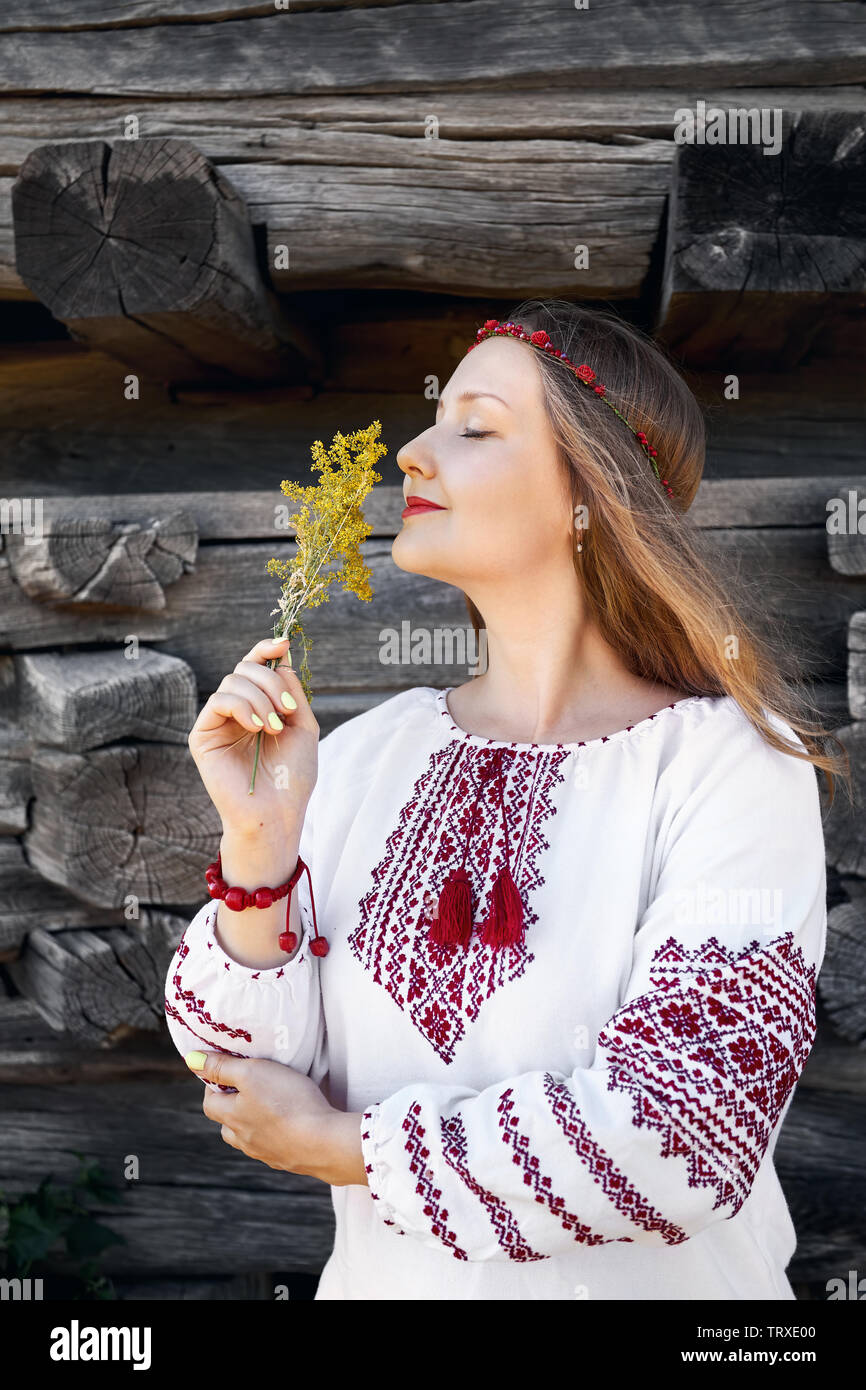Portrait von schönen Mädchen mit langen Haaren in weißen ethnischen Shirt mit Blumenstrauß. Traditionelles Dorf in Osteuropa. Nationale Architektur muse Stockfoto