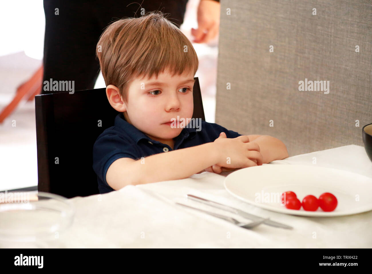 Hungriges Kind im Stuhl sitzend am Tisch in der Küche und wartet auf Essen und Mittagessen. Süße kleine Junge ist neugierig, was er für die Mahlzeit erhalten. Stockfoto