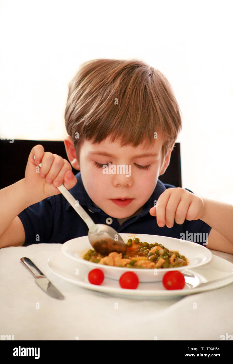 Hungriges Kind im Stuhl sitzend am Tisch in der Küche und essen mit Löffel, gekochte Erbsen mit Tomaten. Kinder essen und gesunde Ernährung Lebensmittel Konzept. Stockfoto