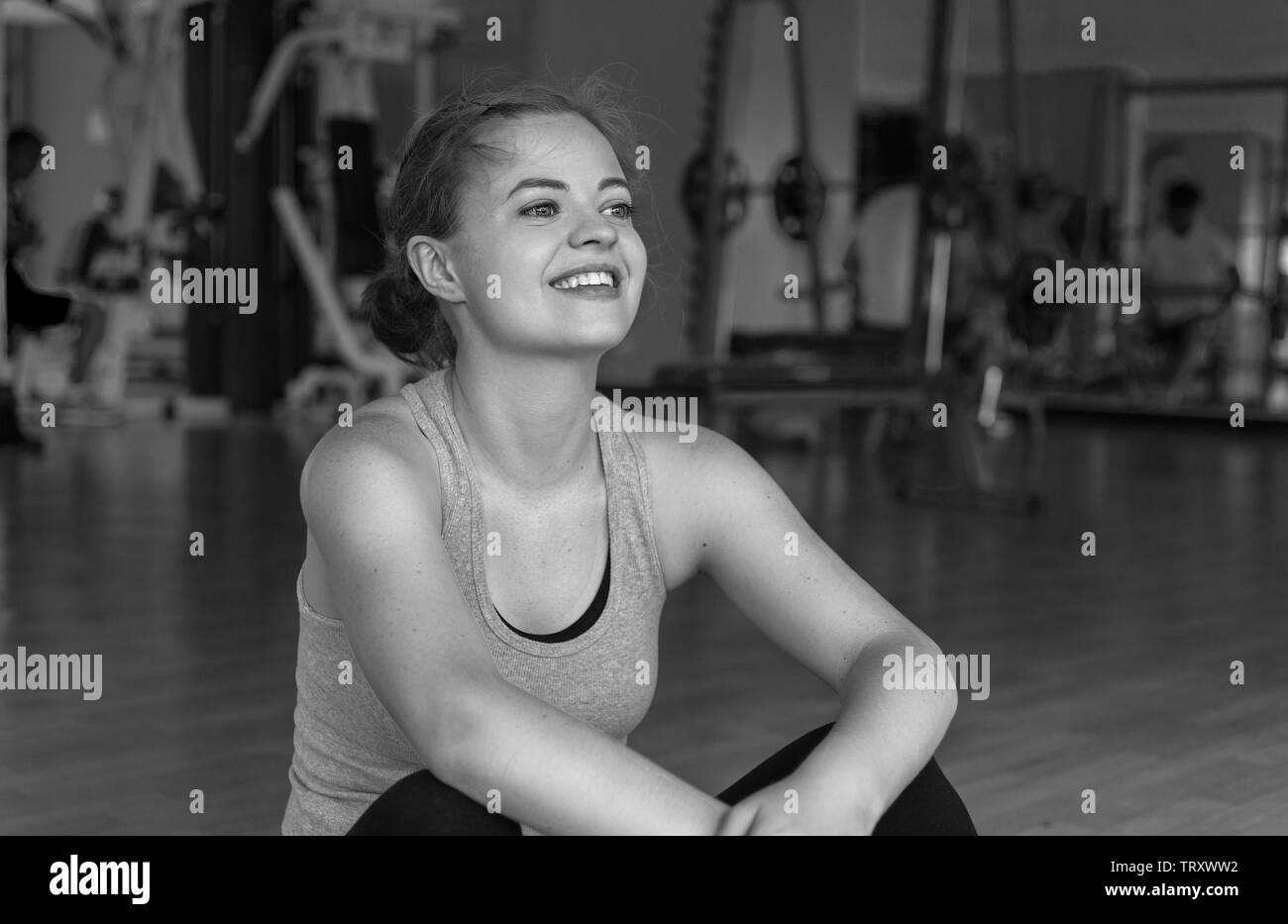 Glückliche Junge kaukasier Frau sitzt auf einer Matte im Fitnessstudio Lächeln, Schwarz und Weiß Stockfoto