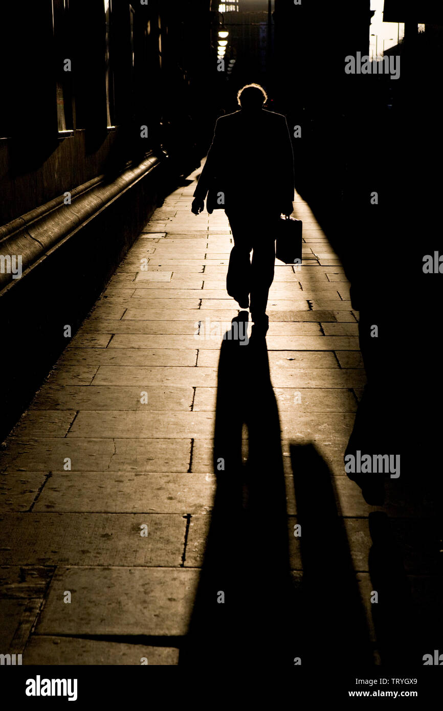 Ein Mann geht in goldenem Winterlicht, umgeben von schweren dunklen Schatten, auf einem Bürgersteig entlang Stockfoto