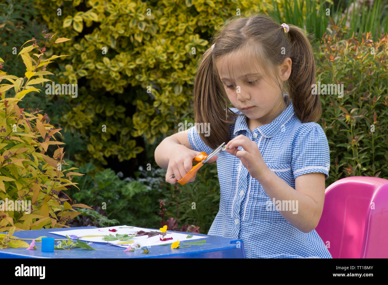 Acht Jahre alten Mädchen in Schuluniform Kleid in den Garten, macht ein Bild von Blumen und Blättern auf Papier geklebt, Natur, Kunst, Handwerk, mit einer Schere Stockfoto