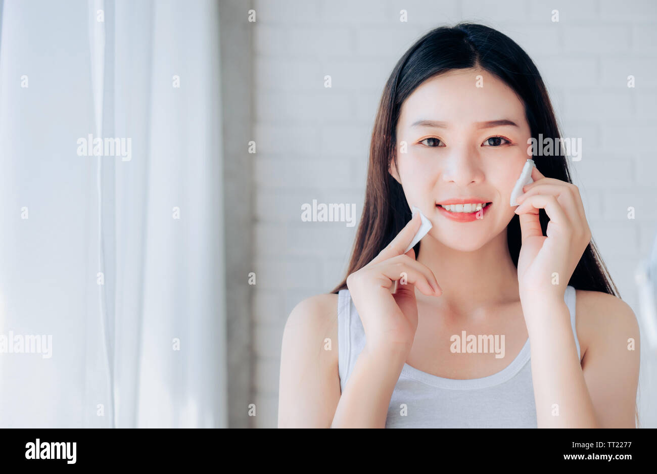 Junge Schönheit asiatische Frau Reinigung Gesicht mit Baumwolle mit Hautpflege und Lächeln Morgen im Weißen Schlafzimmer, Kopieren. Stockfoto