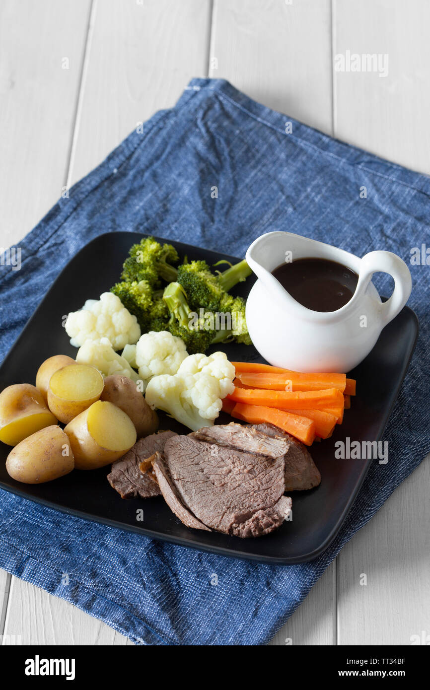 Lammbraten Sonntag Mittagessen Abendessen mit Gemüse auf einem tjeukemeer schwarze Platte, mit Soße Krug, auf einem blauen Küchenhandtuch. Weiß Holz Hintergrund Stockfoto