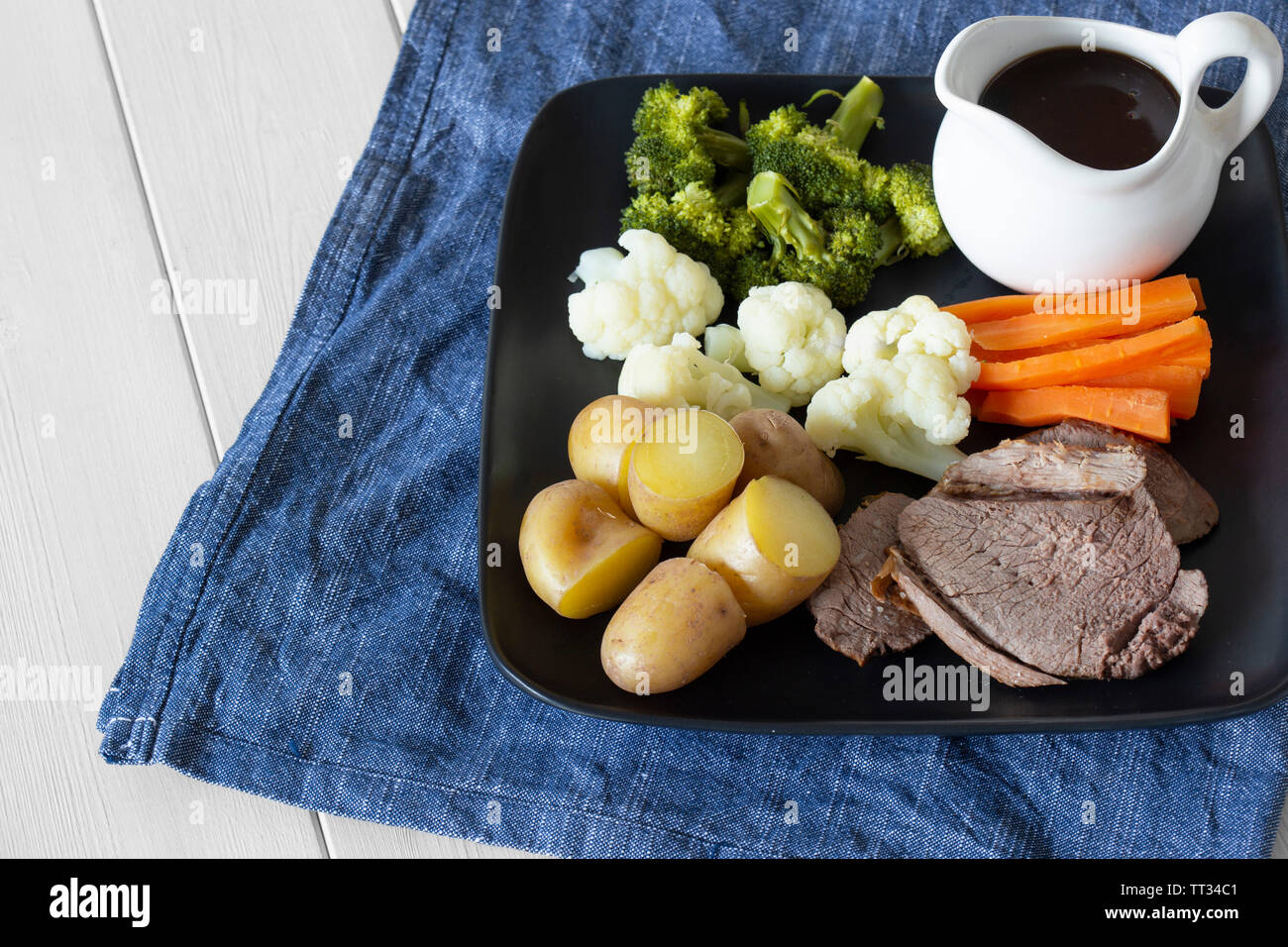 Lammbraten Sonntag Mittagessen Abendessen mit Gemüse auf einem tjeukemeer schwarze Platte, mit Soße Krug, auf einem blauen Küchenhandtuch. Weiß Holz Hintergrund Stockfoto