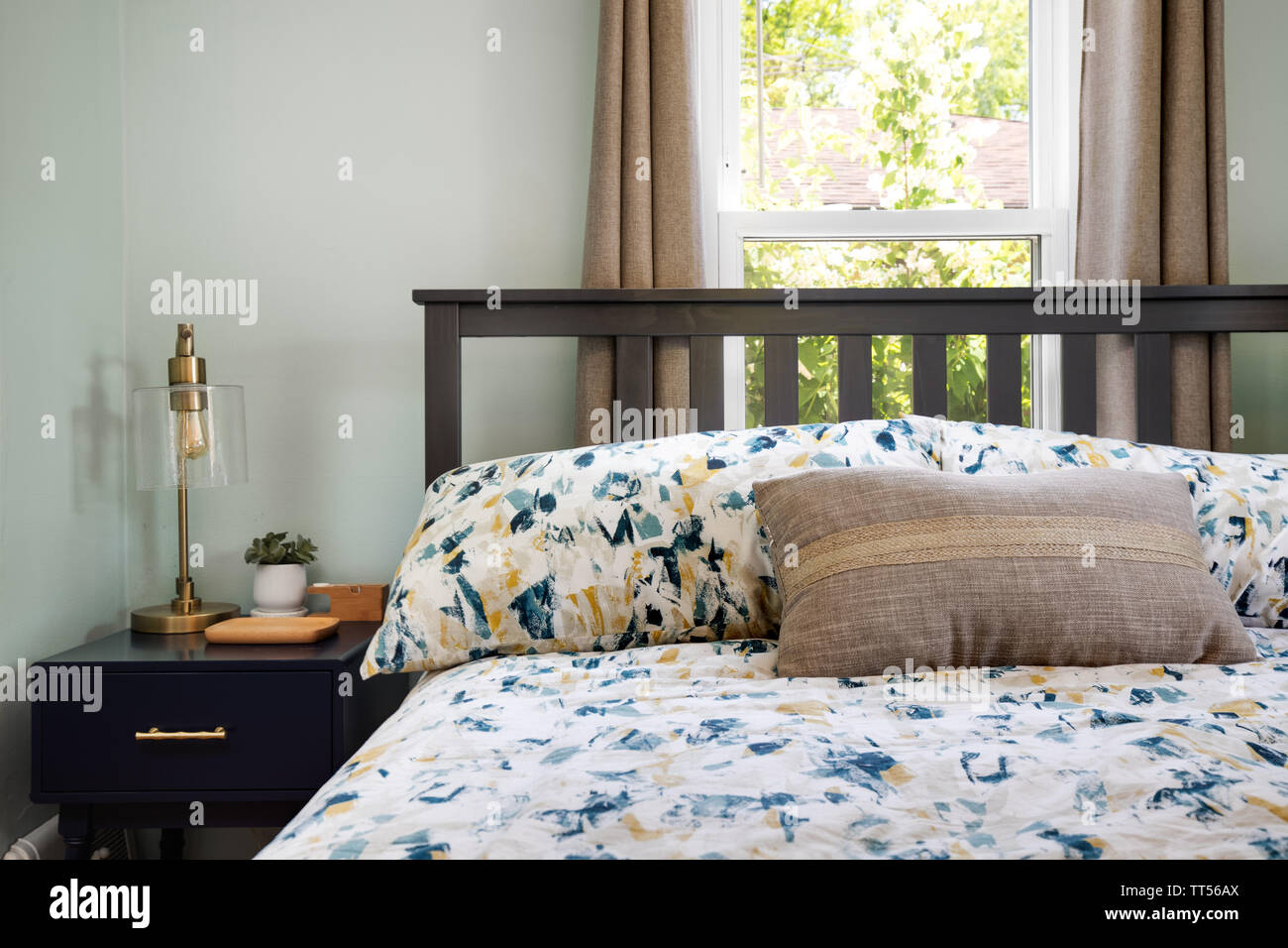 Ein komfortables Zimmer mit einem Bett vor einem Fenster an einem sonnigen  Tag und ein Nachttisch mit einigen Objekten auf Stockfotografie - Alamy