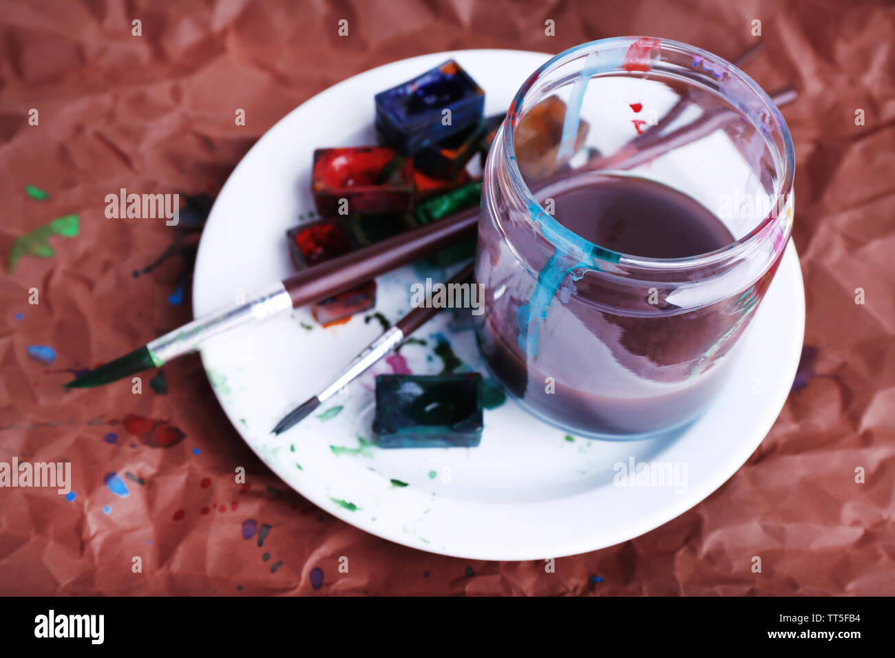 Aquarellfarbe Würfel mit Pinsel und Glas mit Wasser an der Platte auf braunem Papier Hintergrund Stockfoto