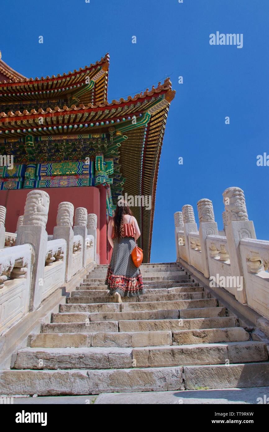 Eine langhaarige, brünett, cauacasian weiblichen touristische Wanderungen bis die Marmorstufen vor einer traditionellen chinesischen Gebäude. Hell Gebäude con lackiert Stockfoto