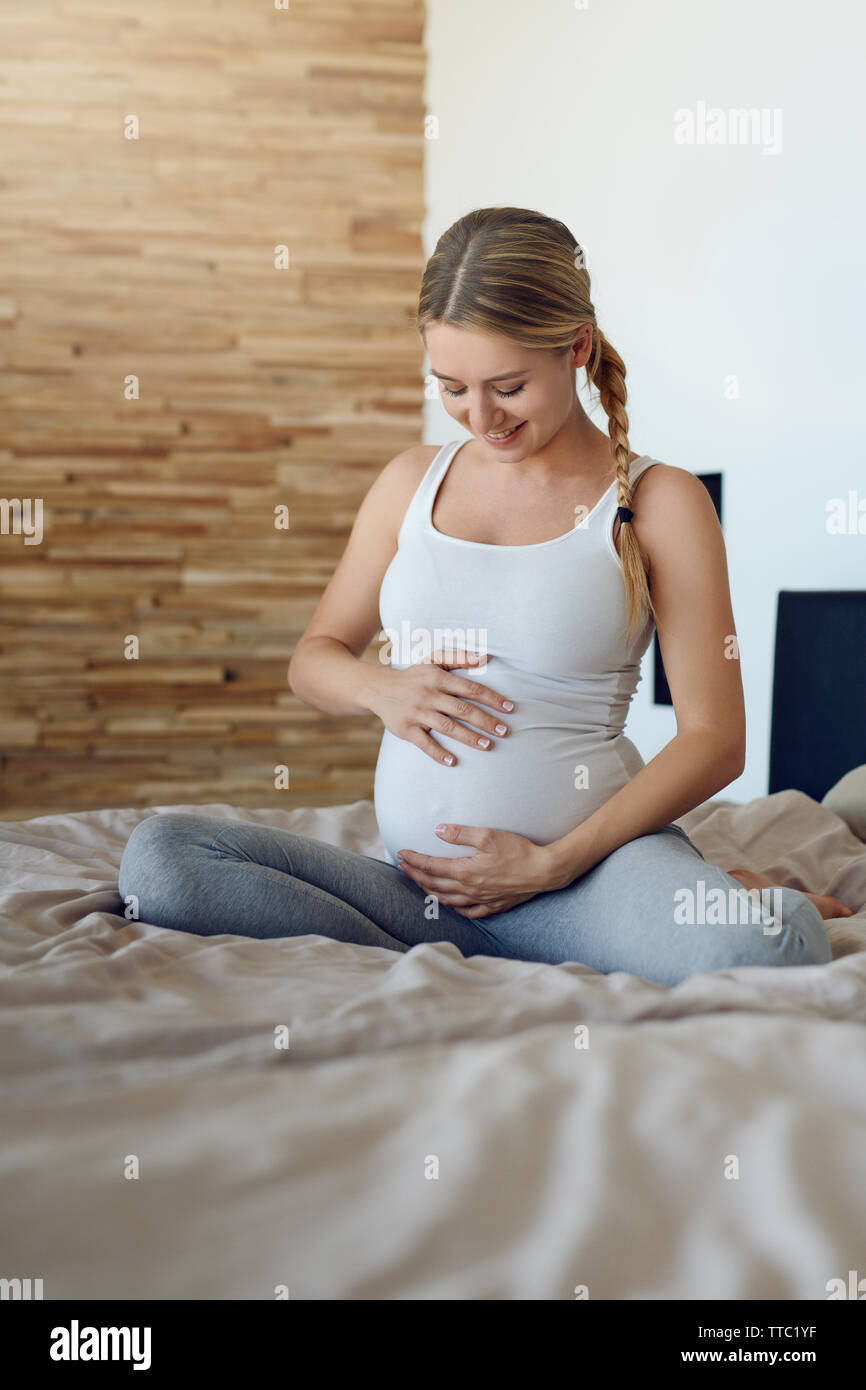 Glückliche junge schwangere Frau Verklebung mit ihrem ungeborenen Kind sitzen auf einem Bett mit Ihrem Babybauch mit ihren Händen nach unten schauen. Stockfoto