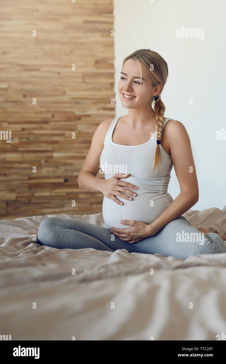 Glückliche junge schwangere Frau Verklebung mit ihrem ungeborenen Kind sitzen auf einem Bett mit Ihrem Babybauch mit den Händen seitlich mit einem zarten Lächeln o Stockfoto