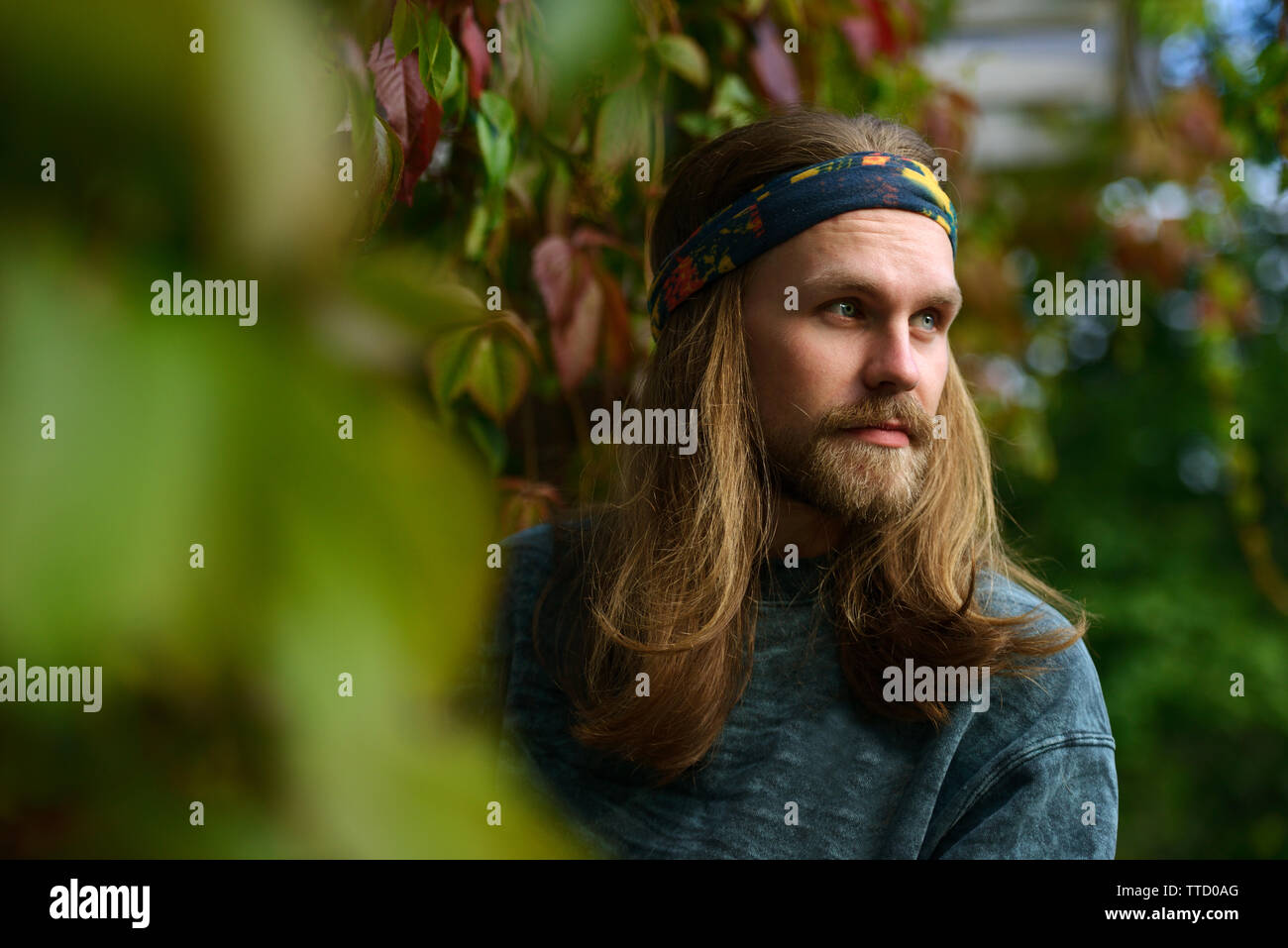 Porträt eines jungen Mannes mit langen Haaren und Bart in ein bandana vor dem hintergrund der grünen Pflanzen Stockfoto