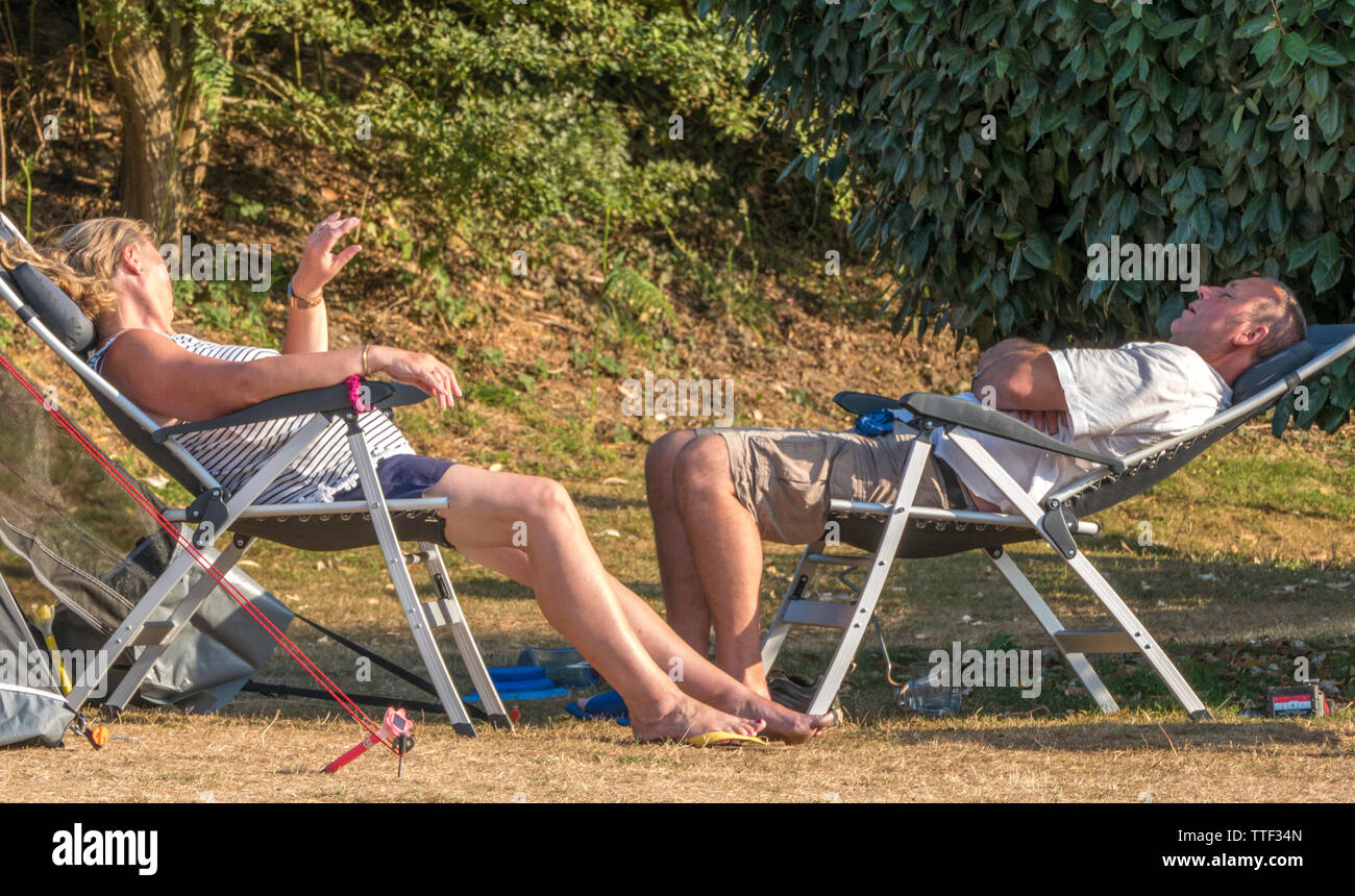 Ein Paar mittleren Alters, die außerhalb in der heißen Sonne auf den Liegestühlen, neben ihr Zelt auf einem Campingplatz in England, Großbritannien. Stockfoto