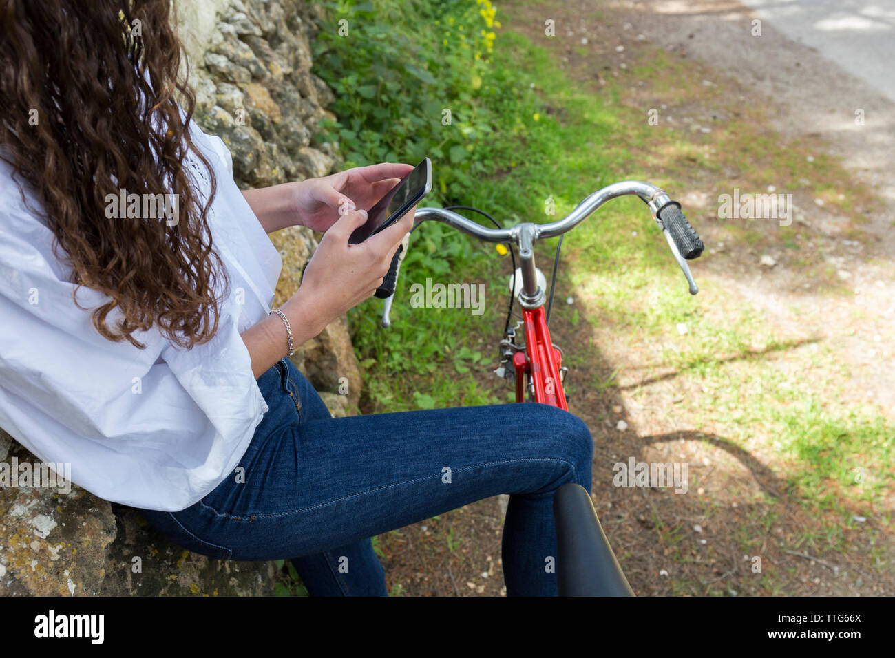 Mittelteil der Frau mit Handy beim Stehen mit dem Fahrrad durch die Mauer aus Stein Stockfoto