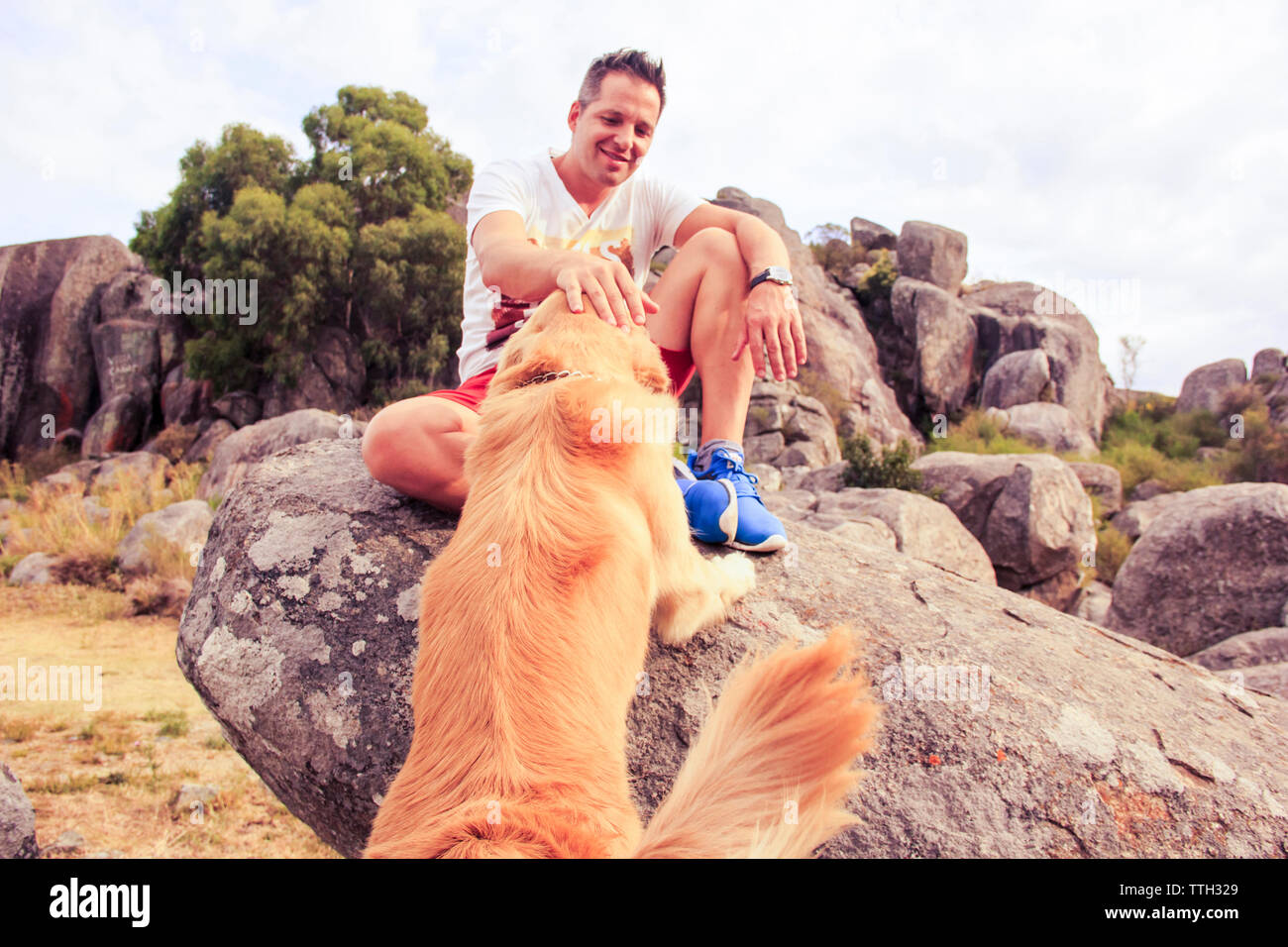 Lächelnd Mitte der erwachsene Mann petting Hund sitzend auf Rock Stockfoto