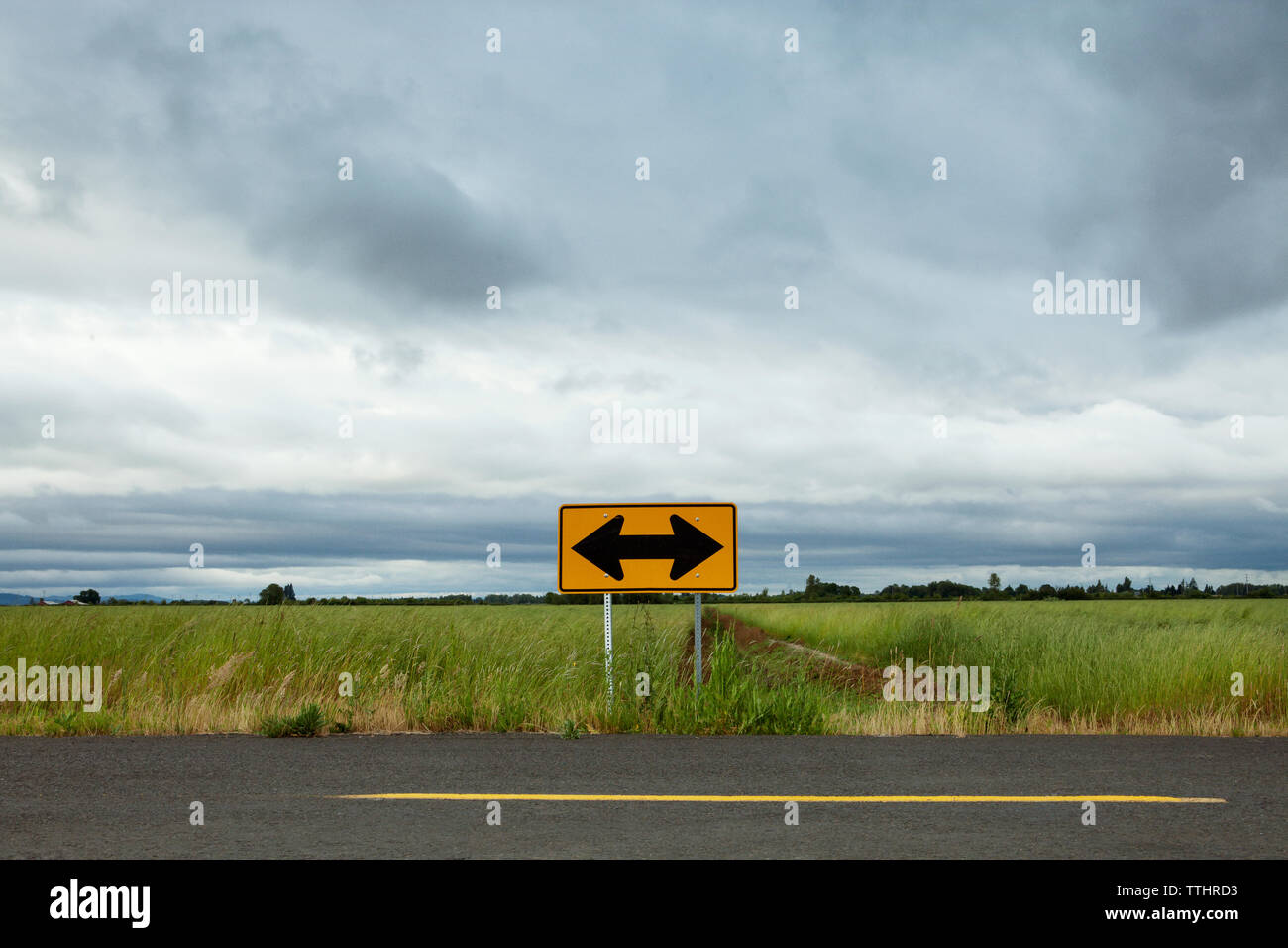 Pfeilsymbol auf leere Straße durch Feld gegen bewölkter Himmel Stockfoto