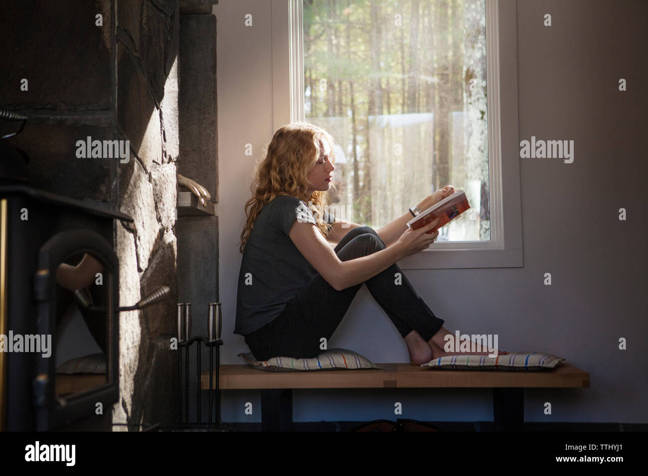 Frau mit Buch beim Sitzen durch Fenster zu Hause Stockfoto