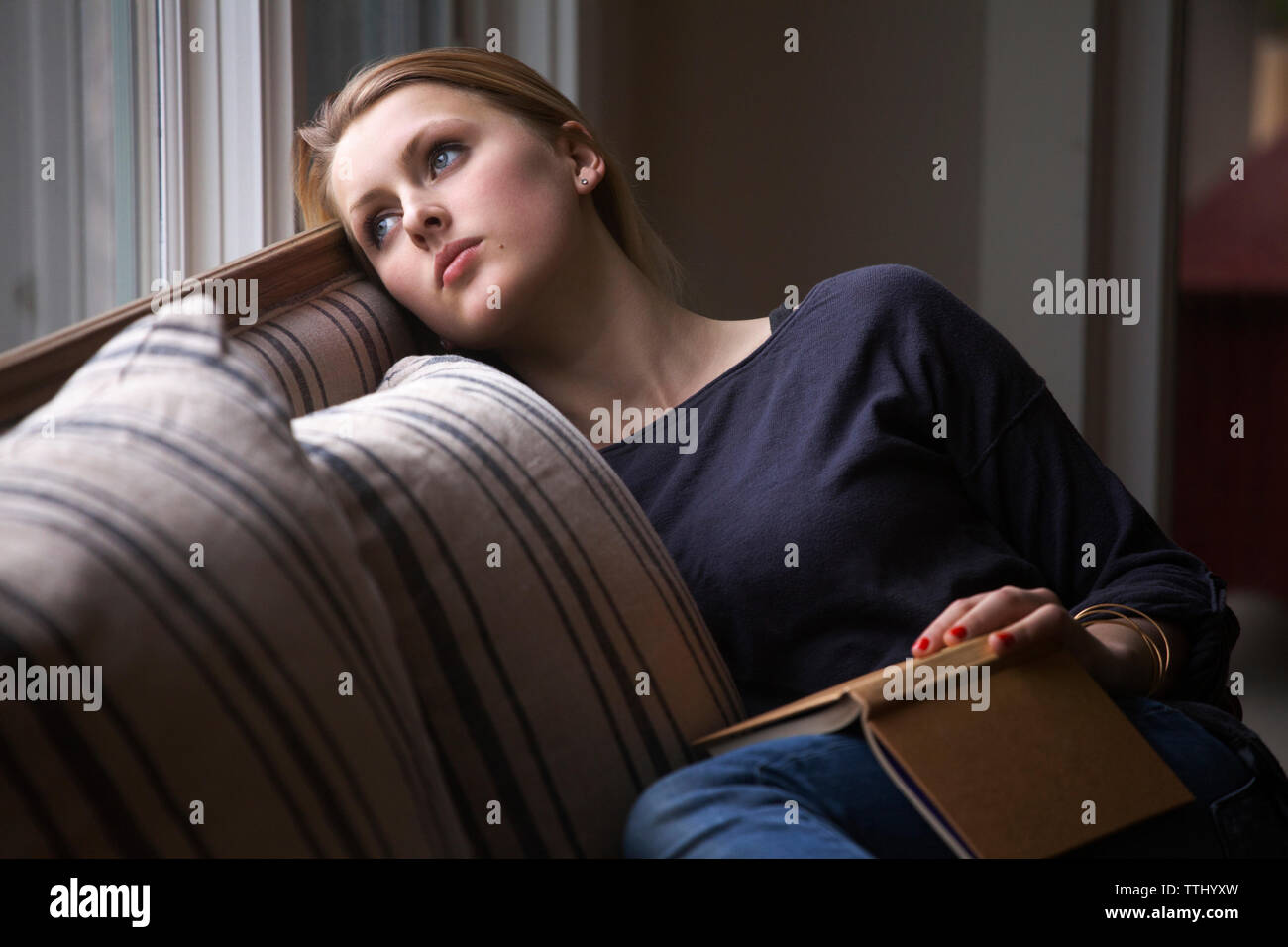 Nachdenkliches Mädchen mit Buch in der Hand weg schauen, und entspannen Sie auf dem Sofa zu Hause Stockfoto